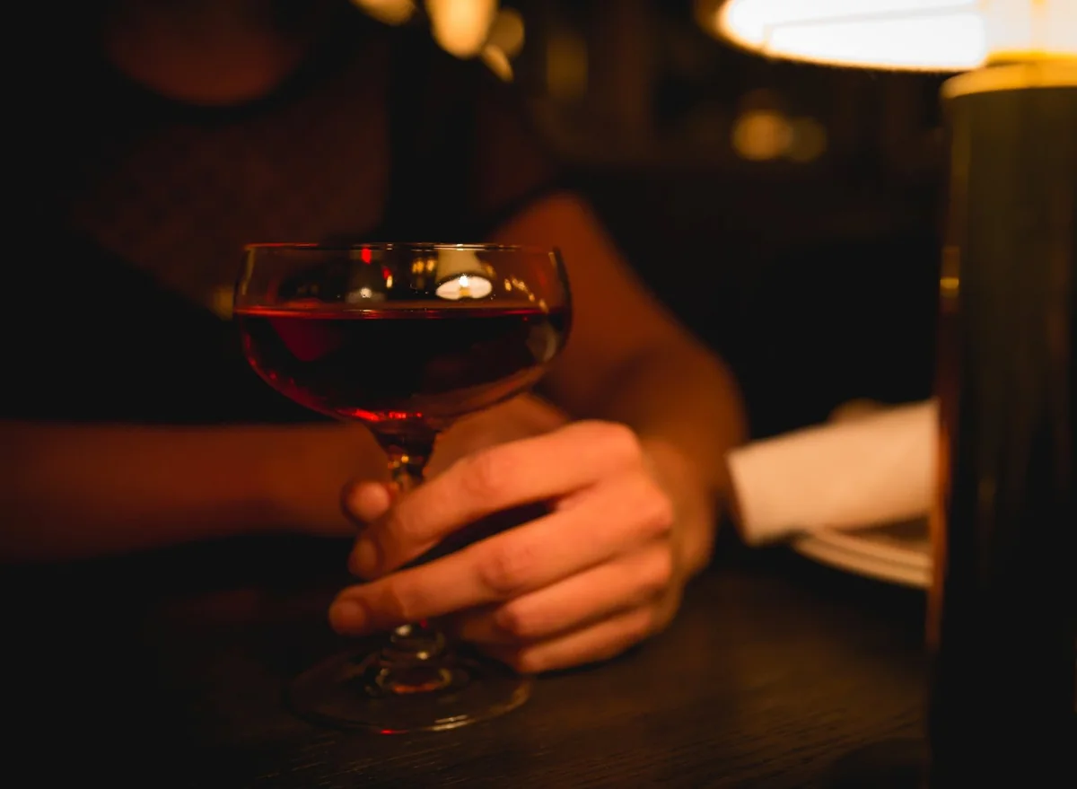 A hand resting on a dark cocktail in a coupe glass under a table lamp.