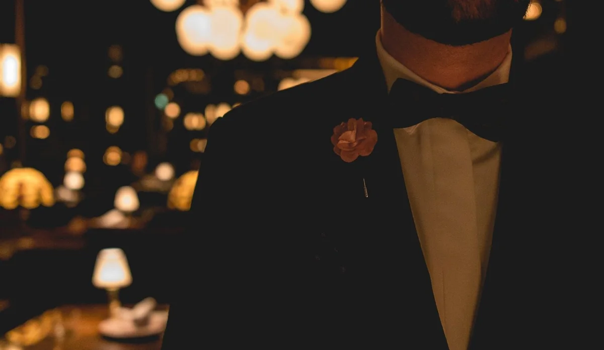A host in a dark dinner jacket and bow tie with a pink boutonniere, the dining room's lamps glowing out of focus behind him.