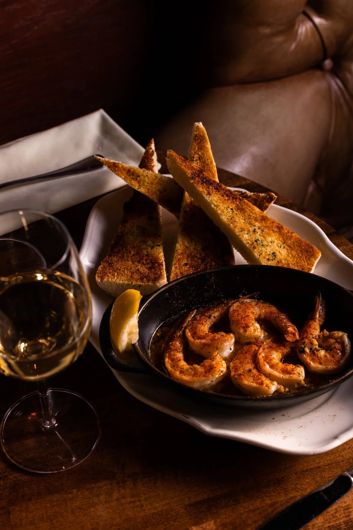Shrimp in a cast-iron dish with garlic toast points, a lemon wedge and a glass of white wine.