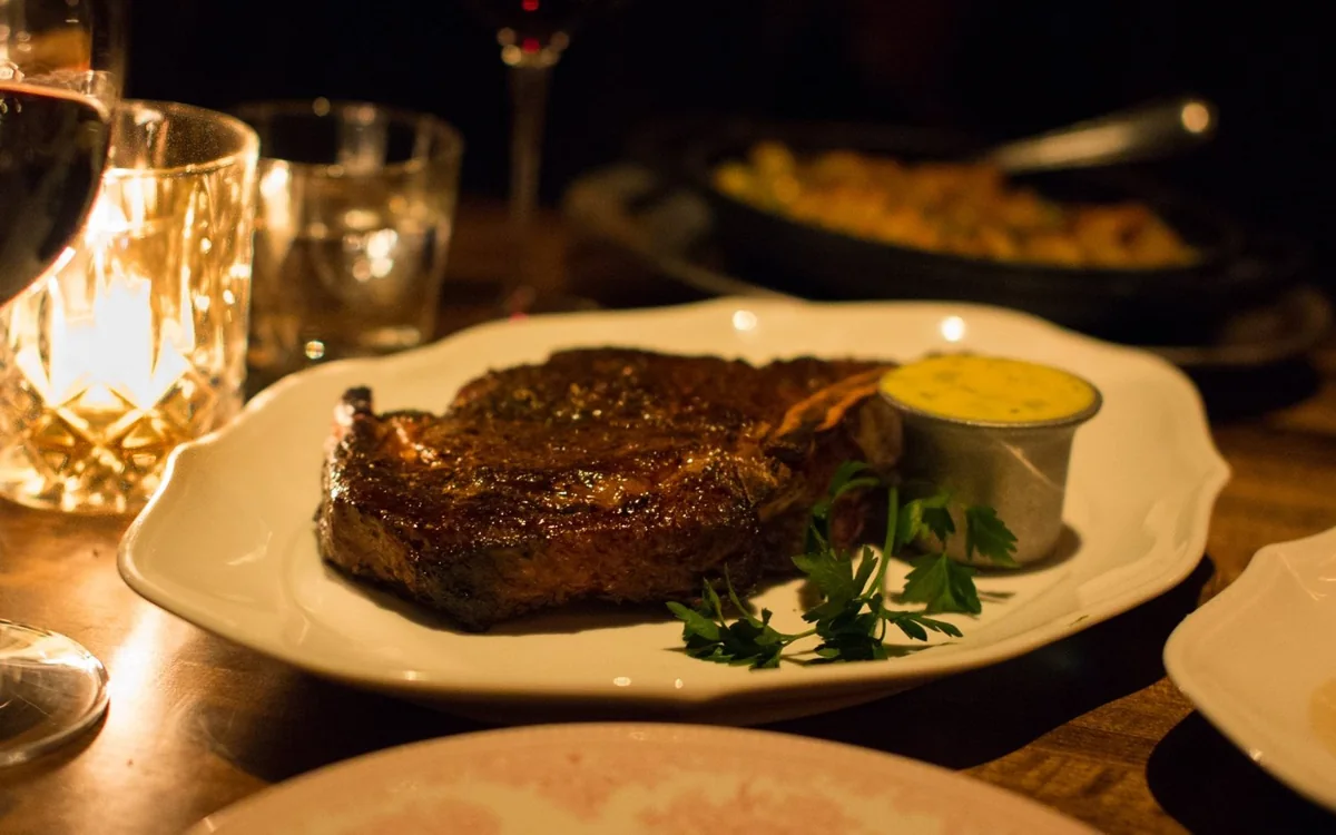 A seared steak on the bone with a cup of butter sauce and parsley on a white plate.