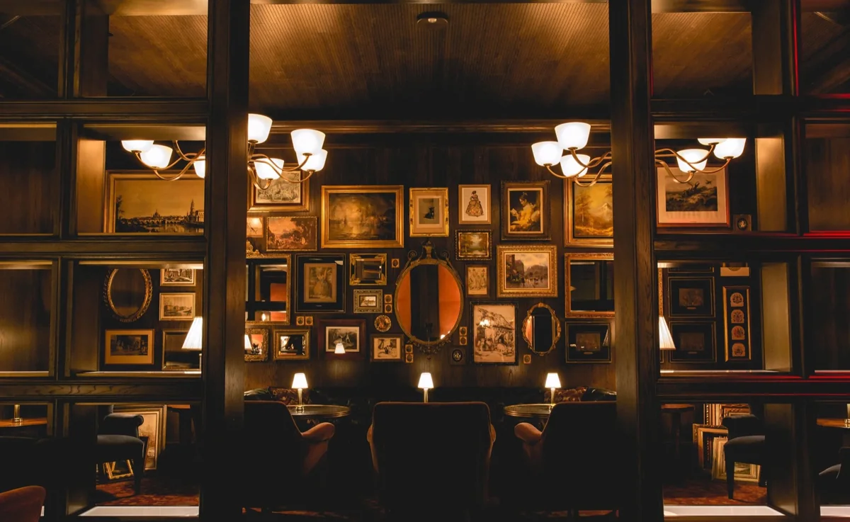 Bavette's Las Vegas: a dark wood-panelled room with a wall of gilt-framed paintings and an oval mirror, two brass chandeliers, small table lamps and upholstered armchairs.